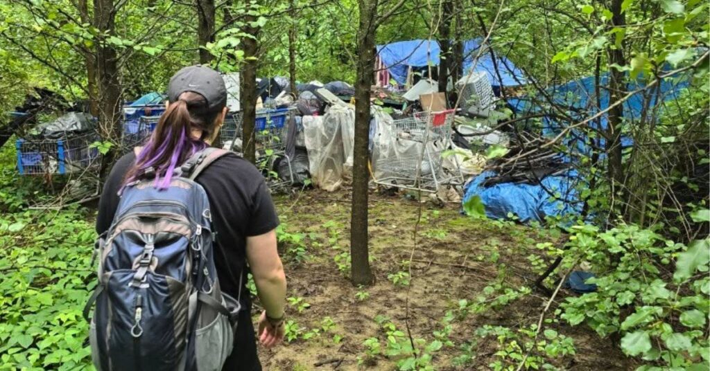 Outreach worker approaching a wooded encampment with belongings and makeshift shelters.